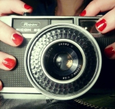 Cámara de fotos cogida por una mano con uñas de color rojo.