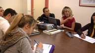 Imagen de una reunión de empresa en la que aparecen varias personas sentadas alrededor de una mesa. Todos están tomando apuntes a excepción de una mujer que, junto a un proyector, explica algo a sus compañeros.
