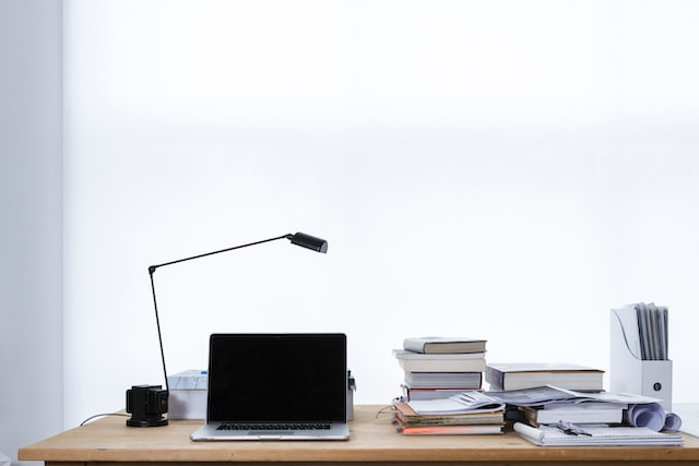 Un portátil encima de una mesa de madera con un flexo y libros alrededor